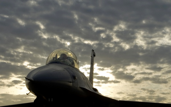 A military fighter jet, consistent with an F-16 Fighting Falcon, is photographed head-on in dramatic silhouette against a sunset or sunrise sky with scattered clouds. The aircraft is stationary on a tarmac or flight line, with a pilot visible in the cockpit. The backlighting creates a striking contrast between the dark aircraft and the golden, cloud-streaked sky.