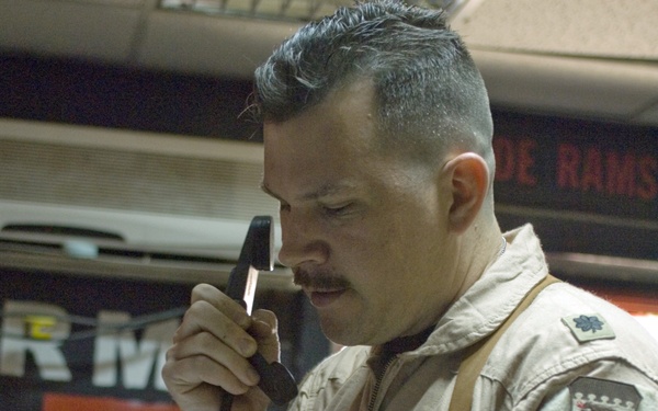 A male military officer in a tan/desert flight suit is speaking on a corded telephone handset while reading from a document. He wears rank insignia consistent with a Lieutenant Colonel (oak leaf cluster) on his shoulder, along with unit patches including what appears to be an Air Expeditionary Wing patch. The setting appears to be a military operations or command center.