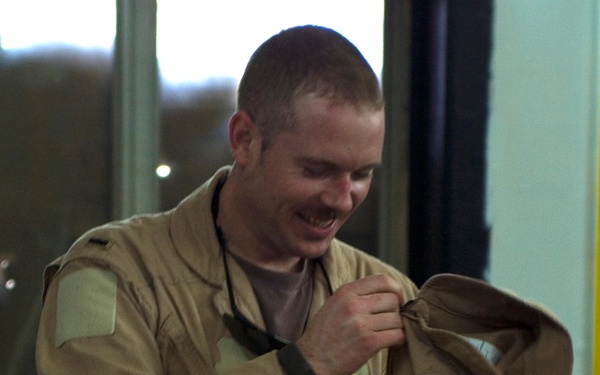 A young man with short hair and a mustache is smiling while examining or handling a tan/khaki military-style flight suit or uniform jacket. He appears to be indoors, possibly inspecting insignia or patches on the garment. The setting appears to be a casual indoor environment with windows visible in the background.