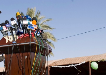 A U.S. Army officer in ACU (Army Combat Uniform) is delivering a speech at a podium laden with numerous media microphones bearing Arabic-language broadcast network logos. The event appears to be an outdoor press conference or official ceremony in what appears to be a Middle Eastern setting, with a covered seating area visible in the background hosting civilian and military attendees. The presence of multiple Arabic-language media outlets and the desert environment suggest this is a military public affairs event likely in Iraq or Afghanistan.