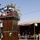 A U.S. Army officer in ACU (Army Combat Uniform) is delivering a speech at a podium laden with numerous media microphones bearing Arabic-language broadcast network logos. The event appears to be an outdoor press conference or official ceremony in what appears to be a Middle Eastern setting, with a covered seating area visible in the background hosting civilian and military attendees. The presence of multiple Arabic-language media outlets and the desert environment suggest this is a military public affairs event likely in Iraq or Afghanistan.