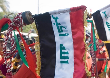 The image shows a military or ceremonial band formation, with personnel dressed in red and black uniforms. Prominent in the foreground are decorative banner flags attached to instruments, displaying the colors of the Iraqi flag (black, white, and red stripes) with green Arabic script. The scene appears to be an outdoor ceremonial parade or official event.