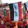 The image shows a military or ceremonial band formation, with personnel dressed in red and black uniforms. Prominent in the foreground are decorative banner flags attached to instruments, displaying the colors of the Iraqi flag (black, white, and red stripes) with green Arabic script. The scene appears to be an outdoor ceremonial parade or official event.