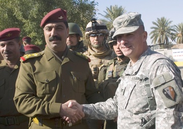A U.S. Army officer in ACU (Army Combat Uniform) is shaking hands with a senior officer in an olive-drab uniform wearing a maroon beret, likely an Iraqi Army general based on the star insignia on his shoulder boards. Several other military personnel are visible in the background, including soldiers in full combat gear and others wearing maroon berets. The setting appears to be an outdoor military or official meeting location with palm trees visible in the background.