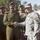 A U.S. Army officer in ACU (Army Combat Uniform) is shaking hands with a senior officer in an olive-drab uniform wearing a maroon beret, likely an Iraqi Army general based on the star insignia on his shoulder boards. Several other military personnel are visible in the background, including soldiers in full combat gear and others wearing maroon berets. The setting appears to be an outdoor military or official meeting location with palm trees visible in the background.