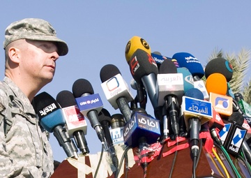 A U.S. Army soldier in ACU (Army Combat Uniform) stands at a podium covered with numerous media microphones from various Arabic-language news organizations. The press conference setting, indicated by the large cluster of microphones with Arabic text logos, suggests a significant military briefing or announcement. The outdoor setting with palm trees in the background indicates a Middle Eastern location.