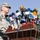A U.S. Army soldier in ACU (Army Combat Uniform) stands at a podium covered with numerous media microphones from various Arabic-language news organizations. The press conference setting, indicated by the large cluster of microphones with Arabic text logos, suggests a significant military briefing or announcement. The outdoor setting with palm trees in the background indicates a Middle Eastern location.