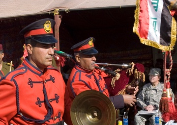 A military marching band in red and black ceremonial uniforms performs at what appears to be an official Iraqi military ceremony or parade. Band members are playing instruments including cymbals and bagpipes, while an Iraqi flag with gold fringe is prominently displayed. A US military soldier in ACU camouflage uniform is seated in the background as an observer or dignitary.