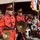 A military marching band in red and black ceremonial uniforms performs at what appears to be an official Iraqi military ceremony or parade. Band members are playing instruments including cymbals and bagpipes, while an Iraqi flag with gold fringe is prominently displayed. A US military soldier in ACU camouflage uniform is seated in the background as an observer or dignitary.
