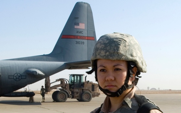 A female U.S. Army soldier stands on an airfield tarmac in full combat gear, holding an M16/M4-style rifle with an optic scope. Behind her is a U.S. Air Force C-130 Hercules transport aircraft bearing the tail markings 'AMC', 'THE ROCK', and serial number '31039', along with ground support equipment. The setting appears to be a forward operating base or military airfield in an arid environment.
