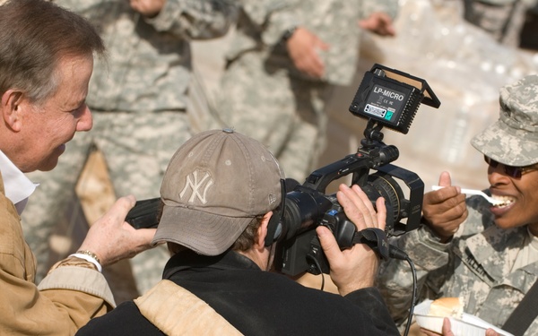 A civilian man in a tan jacket and a videographer wearing a New York Yankees cap are filming or interviewing a female U.S. Army soldier in ACU (Army Combat Uniform) who is smiling and holding a food container. The setting appears to be a military base or forward operating location, with other soldiers visible in the background. The interaction appears to be a media or press visit to a military installation.