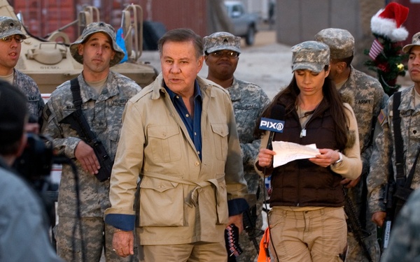 A news crew from PIX11 is conducting a broadcast or interview at what appears to be a military base, surrounded by U.S. Army soldiers in camouflage uniforms. A civilian man in a tan outfit stands centrally while a female reporter holds a PIX11 microphone and reviews notes. A cameraman is visible in the foreground filming the scene.