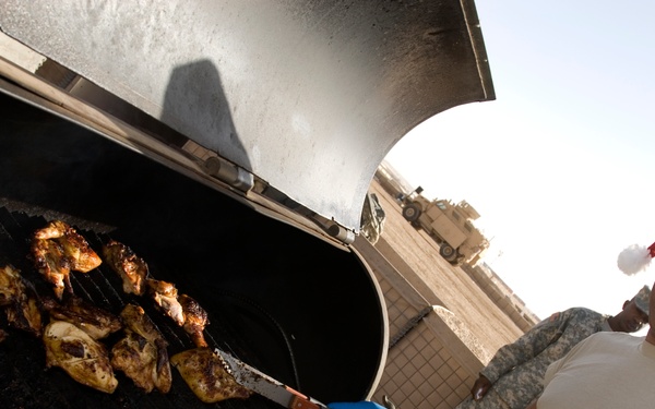 A military service member wearing a Santa hat and ACU (Army Combat Uniform) pants grills what appears to be chicken pieces on a large outdoor barbecue grill. The setting appears to be a forward operating base or military installation, evidenced by military vehicles visible in the background. The scene suggests a holiday celebration, likely Christmas, being observed by deployed troops.