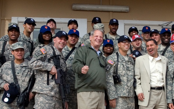 A group photo of approximately 20-25 U.S. Army soldiers in ACU (Army Combat Uniform) camouflage posing with three civilians in front of a building. The soldiers are wearing various New York sports team caps, including New York Yankees (NY) and New York Giants (NY Giants) hats, suggesting a morale or goodwill visit. Several soldiers are carrying M4/M16-style rifles, and the group appears to be in good spirits.