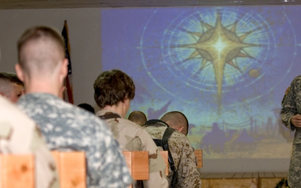 A military chaplain or officer in US Army ACU (Army Combat Uniform) stands at the front of a room addressing seated service members from multiple branches. The setting appears to be a forward operating base chapel or meeting room, with a projected image of a star or compass rose on the screen behind the speaker. Decorative garland and a star ornament suggest this may be a Christmas or holiday religious service.