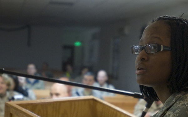 A female soldier in U.S. Army combat uniform (ACU) is speaking at a wooden podium with a microphone stand in front of her. A male soldier stands behind her to the right. The setting appears to be a military chapel or assembly hall with rows of seated personnel in the background.