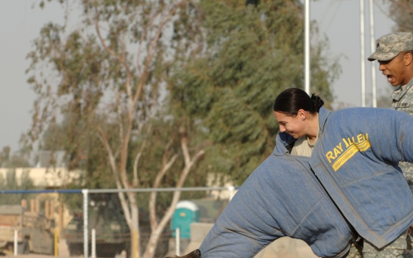Two U.S. Army soldiers in ACU (Army Combat Uniform) are conducting a military working dog (MWD) bite training exercise. One soldier is wearing a 'Ray Allen' brand bite/protection suit and is being engaged by a German Shepherd during a controlled bite drill. A second soldier wearing an 'MP' (Military Police) brassard observes and supervises the exercise.