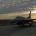 A military fighter jet, consistent with an F-16 Fighting Falcon, is parked on a tarmac at what appears to be a military air base during sunrise or sunset. The aircraft is photographed in dramatic low-light conditions with a golden sky breaking through cloud cover in the background. A large concrete blast wall or revetment structure is visible behind the aircraft.