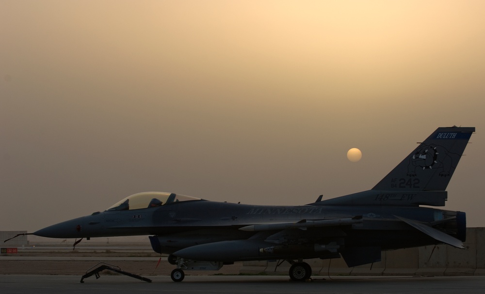 An F-16 Fighting Falcon jet fighter is parked on a tarmac during sunset or sunrise, with a hazy, dust-filtered sky casting an orange glow. The aircraft bears markings indicating it belongs to the Minnesota Air National Guard, based in Duluth. The tail number '242' and the word 'MINNESOTA' are visible on the fuselage.