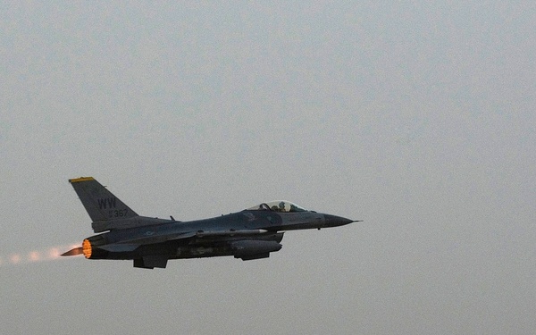 An F-16 Fighting Falcon jet aircraft is captured just after takeoff from a runway, with its afterburner visibly ignited and glowing orange-red. The aircraft bears tail code 'WW' and a partial serial number appearing to read '352', indicating it belongs to a unit using the 'WW' tail code designation. The image was taken in hazy or dusty low-light conditions, likely at dusk or dawn.