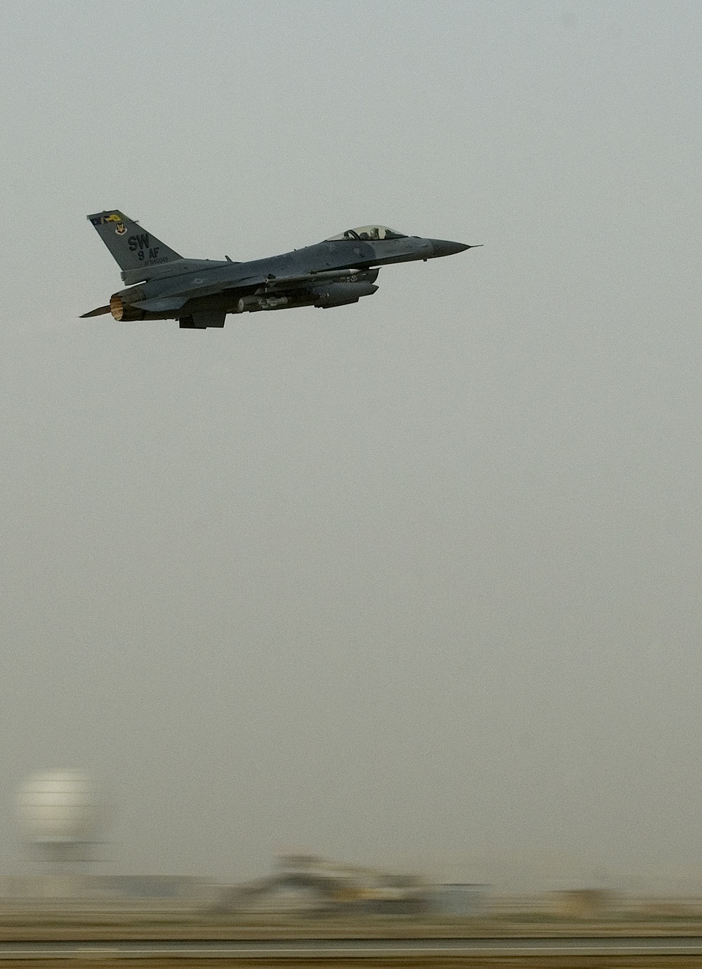 A U.S. Air Force F-16 Fighting Falcon is captured in flight shortly after takeoff, climbing steeply against an overcast, hazy sky. The aircraft bears tail markings 'SW' and '9 AF', indicating assignment to the 20th Fighter Wing (Shaw Air Force Base) under 9th Air Force. The desert terrain and atmospheric haze suggest a Middle Eastern operating environment.