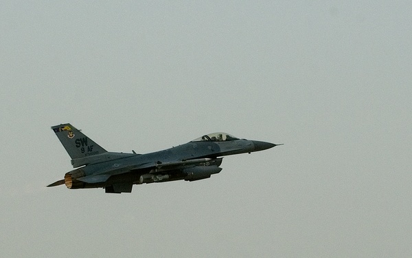 A U.S. Air Force F-16 Fighting Falcon is captured in flight shortly after takeoff, climbing steeply against an overcast, hazy sky. The aircraft bears tail markings 'SW' and '9 AF', indicating assignment to the 20th Fighter Wing (Shaw Air Force Base) under 9th Air Force. The desert terrain and atmospheric haze suggest a Middle Eastern operating environment.