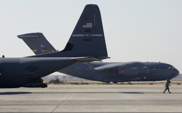 The image shows two U.S. Air Force C-130 Hercules transport aircraft on a tarmac in an arid environment. The foreground aircraft displays tail markings identifying it as belonging to AMC (Air Mobility Command), nicknamed 'The Rock,' with serial number 64631. A single airman is visible walking on the tarmac between the two aircraft.