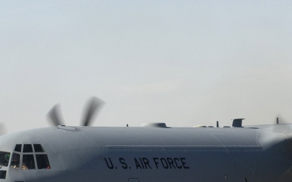A U.S. Air Force C-130 Hercules military transport aircraft is taxiing on a tarmac with all four turboprop engines running, evidenced by the motion blur on the propellers. The aircraft bears the markings '4631' and '463 AG' on its fuselage, along with the U.S. Air Force shield insignia. The setting appears to be a desert or arid-region airfield based on the hazy sky and pale concrete surface.