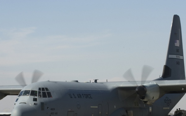 A U.S. Air Force C-130 Hercules military transport aircraft is positioned on a tarmac with all four turboprop engines running, indicated by the motion blur on the propellers. Ground support vehicles and equipment are visible to the right of the aircraft. At least one additional C-130 aircraft is visible in the background to the left.