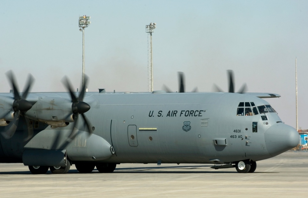A U.S. Air Force C-130 Hercules military transport aircraft is parked on a tarmac, with its four turboprop engines running as indicated by the motion-blurred propellers. The aircraft bears the markings '4631' and '463 AG' on its fuselage, along with the U.S. Air Force shield insignia. The setting appears to be a forward operating base or military airfield in an arid environment.