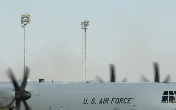 A U.S. Air Force C-130 Hercules military transport aircraft is parked on a tarmac, with its four turboprop engines running as indicated by the motion-blurred propellers. The aircraft bears the markings '4631' and '463 AG' on its fuselage, along with the U.S. Air Force shield insignia. The setting appears to be a forward operating base or military airfield in an arid environment.