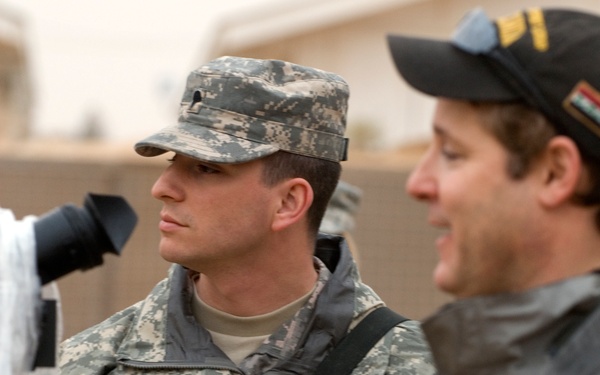 Two men are visible in the foreground, one wearing US Army ACU (Army Combat Uniform) with a matching patrol cap, and another in civilian clothing with a dark baseball cap. A boom microphone or similar audio/video equipment is visible in the frame, suggesting a media or interview setting. The background appears to be an outdoor or semi-outdoor environment with neutral-colored walls.