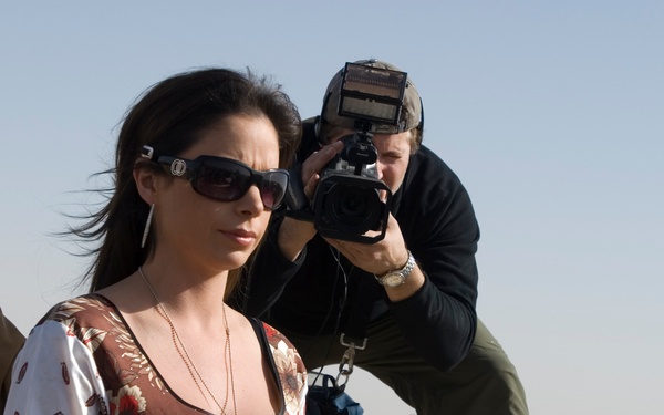 A woman wearing sunglasses and a floral dress stands near a large green plastic container in an arid, desert-like environment. Behind her, a male cameraman crouches while filming with a professional video camera. The background suggests a military or forward operating base setting, indicated by earthen barriers and sparse desert terrain.
