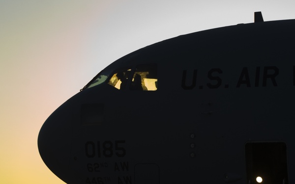 A U.S. Air Force military transport aircraft is photographed on a tarmac at sunrise or sunset, creating a dramatic silhouette effect. The nose section of the aircraft is prominently featured, displaying the tail number '0185' and unit markings '62nd AW' and '446th AW'. The forward entry door is open, and the aircraft is parked on what appears to be a military airfield.