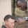 Activities at Al Asad Air Base A military service member in camouflage uniform is giving a haircut to another individual seated in a green barber's cape. The setting appears to be an outdoor or semi-outdoor military environment, likely a forward operating base or field camp. The barber is using electric clippers and a comb to perform the haircut.