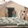 Activities at Al Asad Air Base The image shows U.S. military personnel at a forward operating base, gathered around a 'U.S. BASE-X' tent in a desert environment. A field haircut is being administered outdoors, with one service member cutting another's hair using clippers while the subject sits draped in a green barber's cape. Several other personnel in desert camouflage uniforms are visible in the surrounding area.