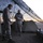 Activities at Al Asad Air Base Two military personnel in desert camouflage uniforms stand beneath a camouflage netting in what appears to be a forward operating base or field camp in an arid desert environment. One individual appears to be examining or handling a small object while the other stands nearby. The setting includes a small dome tent, a makeshift table with supplies, and a large map board visible on the left.