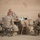 Activities at Al Asad Air Base Five military personnel in desert camouflage uniforms (MARPAT pattern consistent with U.S. Marines) are seated around a folding table in what appears to be a forward operating base or field environment. They appear to be engaged in a briefing or informal meeting, with documents and supplies visible on the table. The setting is outdoors with a tan canvas or tarp backdrop.