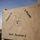 Activities at Al Asad Air Base A hand-painted wooden sign marks a military forward operating base called 'Camp Reasoner II,' belonging to Charlie Company, 1st Reconnaissance Battalion. The sign features a skull-and-crossbones style emblem with crossed weapons, consistent with reconnaissance unit insignia. The sign is mounted on wooden posts in an arid, desert-like environment.