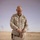 Activities at Al Asad Air Base A U.S. Marines officer in desert MARPAT camouflage uniform, identified by name tape as 'KELLY,' is kneeling and using a knife or tool on what appears to be a cardboard box or wooden object. The setting appears to be an outdoor desert environment with a white backdrop or tent wall behind him. A military helmet and gear are visible on the surface in front of him.