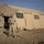 Activities at Al Asad Air Base Two military personnel in desert camouflage uniforms stand outside a large tan military tent in an arid environment. The setting appears to be a forward operating base or military camp, with sandy terrain and sandbags reinforcing the base of the tent. Camouflage netting is visible overhead on the left side of the image.
