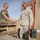 Activities at Al Asad Air Base Two U.S. Marines are shaking hands in what appears to be a forward operating base or desert training/deployment environment. A senior Marine in full desert MARPAT camouflage uniform is greeting a junior Marine wearing a green t-shirt. The setting includes wooden pallets, military supply boxes, ammunition cans, and a shipping container in the background.