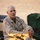 Activities at Al Asad Air Base A U.S. Marine Corps officer in desert MARPAT camouflage uniform is seated on what appears to be a wooden crate or ammunition box in an outdoor desert environment. The name tape 'KELLY' is visible on the uniform, along with 'U.S. MARINES' tape and what appear to be general officer rank insignia on the collar. The individual appears to be engaged in conversation, with folding camp chairs visible in the background.