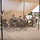Activities at Al Asad Air Base A group of approximately five to six military personnel in desert camouflage uniforms are seated around a folding table under a tan shade canopy in an arid, desert environment. They appear to be engaged in a meeting or meal in what looks like a forward operating base or field camp. Military vehicles, including what appear to be tactical trucks, are visible in the background.