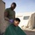 Activities at Al Asad Air Base A military service member in an olive green t-shirt and camouflage trousers is cutting another service member's hair outdoors at what appears to be a forward operating base or military camp. The barber is wearing purple latex gloves and using electric hair clippers, while the seated individual is draped in a green barber's cape. Military-style canvas tents and sandbags are visible in the background.