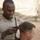 Activities at Al Asad Air Base A U.S. Marine in desert MARPAT camouflage uniform is giving a haircut to another service member using electric clippers in an outdoor field environment. The setting appears to be a forward operating base or field camp, indicated by the tent structures visible in the background. The barber is smiling, suggesting an informal, morale-boosting activity.