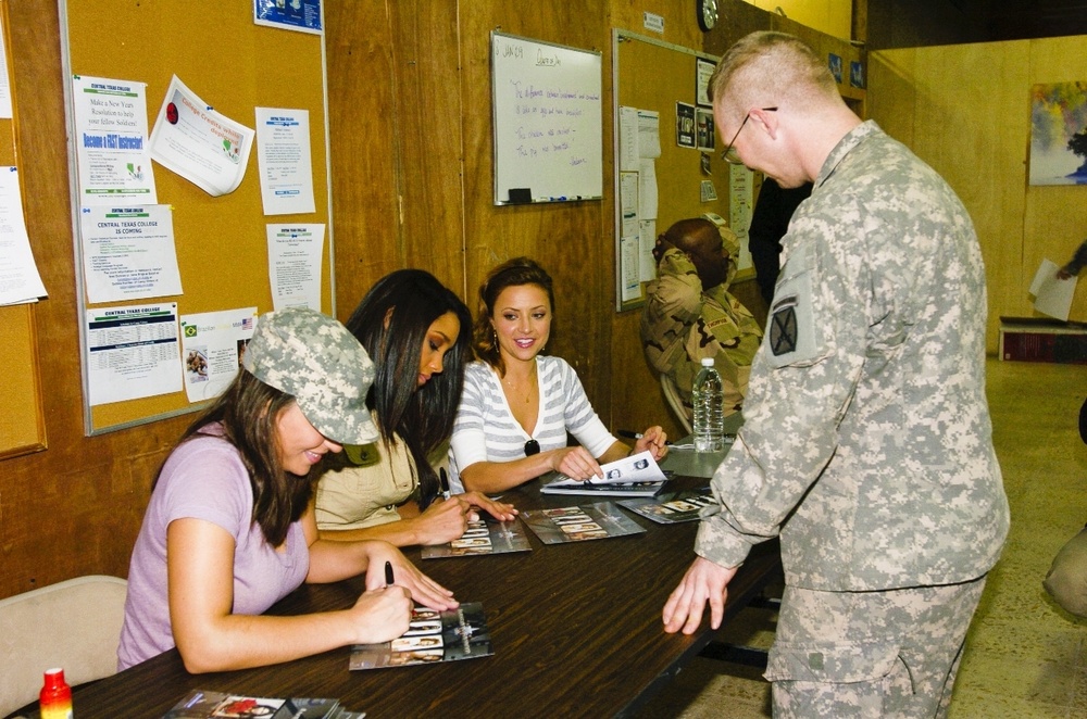A military setting where two civilian women appear to be signing autographs at a table for military personnel. One soldier in Army Combat Uniform (ACU) with a 10th Mountain Division patch stands at the table, while another person wearing a military cap sits nearby. The scene appears to be a USO-style meet-and-greet or morale event at a military installation.