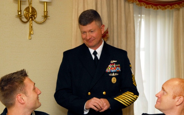 Three U.S. Navy personnel are engaged in conversation at what appears to be a formal dining event or reception. A senior officer in dress blues with extensive gold sleeve stripes and ribbons stands while two enlisted sailors wearing name tags sit at a table set with burgundy napkins and coffee cups. The setting appears to be an upscale venue with ornate curtains and a decorative wall sconce.