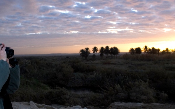 A male individual wearing U.S. Army ACU (Army Combat Uniform) pattern camouflage cap and military-style clothing stands on an elevated rocky terrain, photographing a landscape at either sunrise or sunset. The scene shows a flat, arid environment with scattered low vegetation and a row of palm trees silhouetted against a warm orange horizon. The individual carries a camera and a tan/coyote-colored pouch on his belt.
