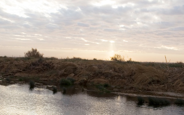 A military service member in camouflage uniform stands on an earthen bank overlooking a calm body of water, likely a river or canal, at dusk or dawn. The landscape is arid and sparse, with dry vegetation and disturbed earth visible on the far bank. The sky is overcast with diffused light breaking through near the horizon.